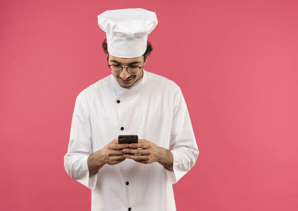 眼镜面带微笑的年轻男厨师穿着厨师制服 戴着眼镜 把电话号码孤立在粉红色的墙上拨号电话微笑