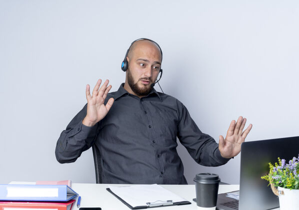 男人给人印象深刻的年轻秃头呼叫中心男子戴着耳机坐在办公桌旁 拿着工作工具看着笔记本电脑 空手对着白色的笔记本电脑做停止手势电话手手势
