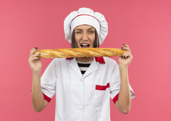 棒自信的年轻女厨师穿着厨师制服拿着面包棒 眨着眼睛孤立在粉红色眨眼烹饪信心