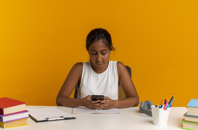 桌子年轻的女学生坐在书桌旁 手里拿着学习用具 电话号码被隔离在橙色的墙上电话工具坐着