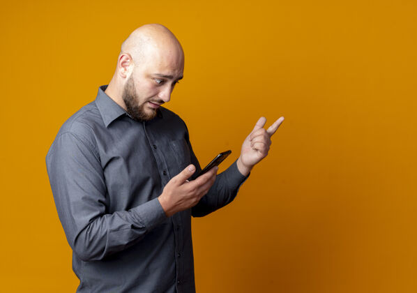 手机印象深刻的年轻秃头呼叫中心的男子站在侧视图举行 并看着手机 指着一边孤立的橙色与复制空间通话橙色中心
