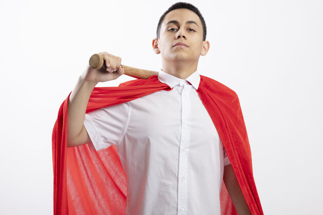 超级英雄自信的年轻超级英雄男孩 穿着红色斗篷 肩上扛着棒球棒 看着白色背景上孤立的摄像机自信年轻肩膀
