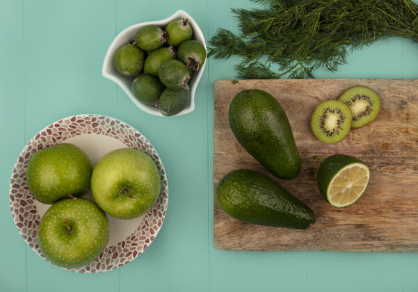 酸橙在蓝色墙上的木制厨房板上 一个碗上放着新鲜的绿色苹果 一个碗上放着菠萝 一个碗上放着鳄梨 酸橙和猕猴桃水果飞珠顶部