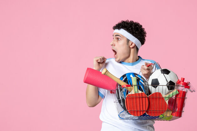 成人正面图穿着运动服的年轻男子 带着装满运动用品的篮子 粉色的墙壁惊喜前面体育