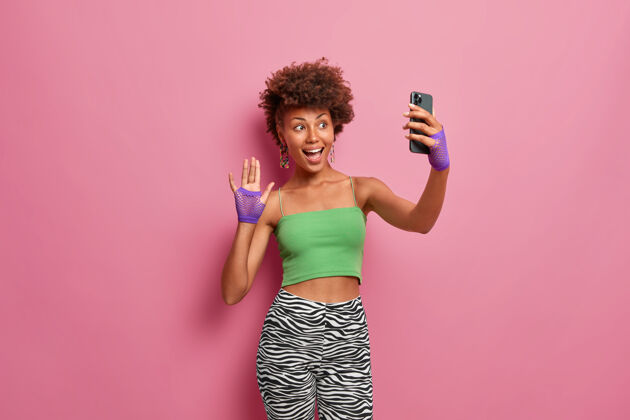 应用程序穿着绿色不规则上衣和紧身裤 戴着运动手套 在智能手机摄像头前挥手的快乐时尚女人 在她的博客上向粉丝致意手机自拍乐趣