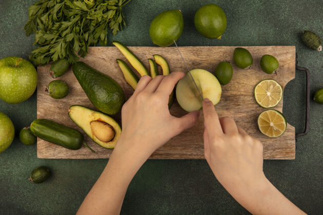 视图女性手用刀在木制厨房板上切新鲜苹果的俯视图 在绿色墙上隔离着酸橙 鳄梨 青苹果和欧芹水果苹果农业