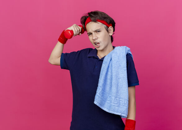 肩膀自信的年轻英俊的运动男孩戴着头带和护腕 肩上戴着牙套和毛巾 举起哑铃 看着深红的墙上孤立的前方运动感觉男孩