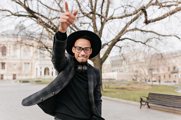 夹克灵感来自身穿灰色夹克的黑人男子在公园挥手致意戴着帽子和眼镜的快乐非洲男模在城市广场休息的户外肖像帽子城市景观黑发