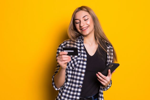 年轻一个快乐的金发女郎展示塑料信用卡的肖像 而使用手机隔离在黄色的墙壁应用程序漂亮信用