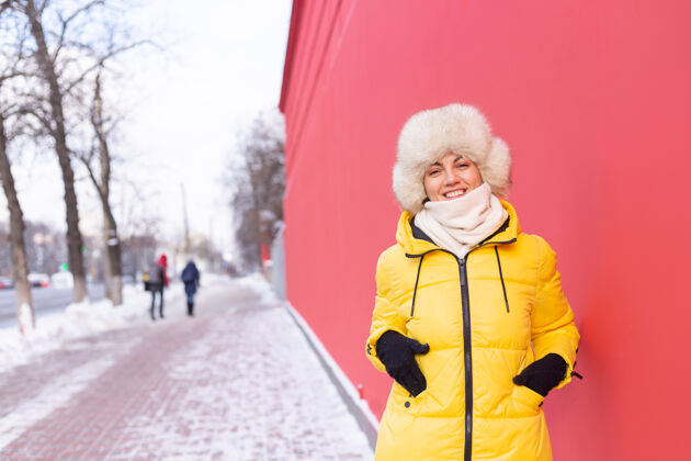 酷在一个冬天阳光明媚的日子里 穿着暖和衣服的红墙背景上的快乐年轻女子衣服成人微笑