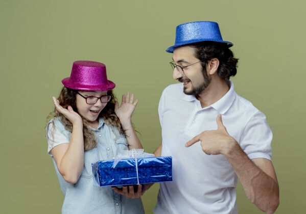 女孩一对戴着粉蓝相间帽子的年轻情侣微笑着把礼盒送给被孤立在橄榄绿上的惊讶女孩年轻惊喜微笑
