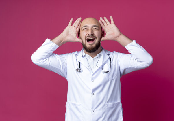 表演快乐的年轻秃头男医生穿着医用长袍和听诊器 在粉红色的墙上显示兔子耳朵的姿势年轻长袍欢乐