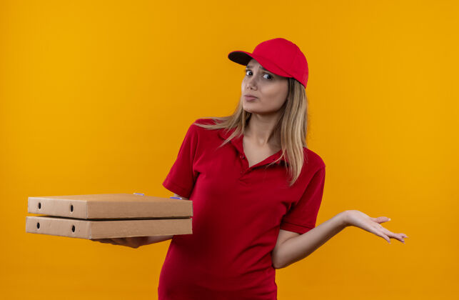 披萨困惑的年轻送货女孩穿着红色制服 戴着帽子 手里拿着披萨盒 摊开手孤立在橙色的墙上蔓延送货年轻