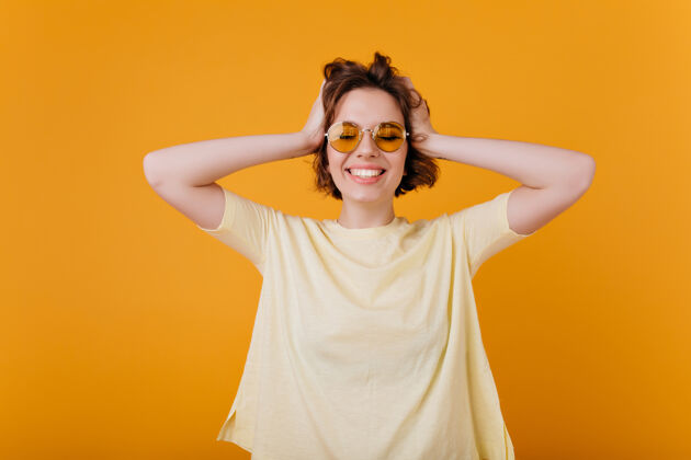 时尚迷人的女士 深色卷发 表达幸福橙色墙壁上隔离着身着黄色服装的幸福的欧洲女孩的室内肖像快乐女人青春