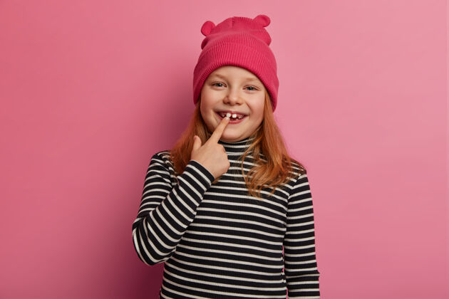 牙医可爱的小姜姑娘露出她第一个成年的两颗牙齿 欢笑着 表达积极的情绪 张嘴 准备做口腔检查 穿着条纹毛衣 戴着粉红色的帽子幼儿园情绪红发