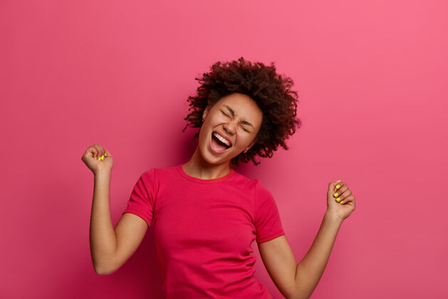 倾斜喜出望外的女人凯旋的照片 拍打拳头 歪着头欢笑 庆祝自己的成功 穿着休闲t恤 获得胜利并实现目标 在粉红色的墙上摆姿势庆祝胜利人类