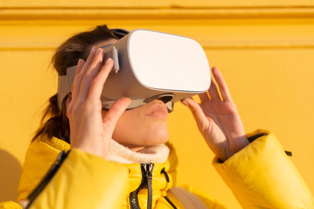 显示一个戴着虚拟现实眼镜的女人的肖像 在冬天的阳光下 穿着暖和的衣服 靠在黄色的墙上女孩虚拟现实游戏现实