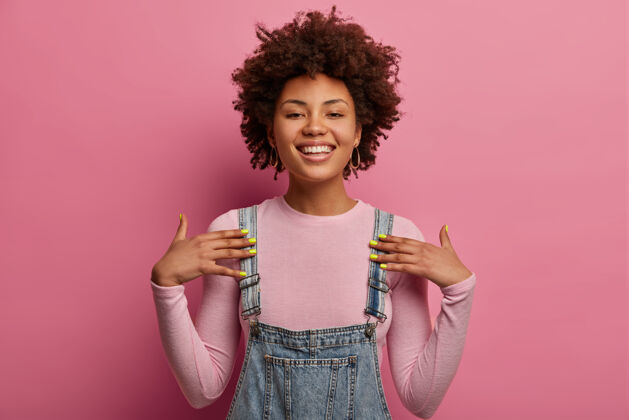 人积极向上的非洲裔年轻女性 自命不凡 面带笑容 精神抖擞 穿着休闲服 对着粉色粉彩墙摆姿势 有自信的表情学生室内美国