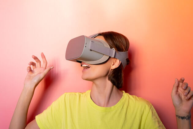 虚拟现实在粉红色橙色背景的工作室里 戴着虚拟现实眼镜的女人的情感肖像智能现实设备