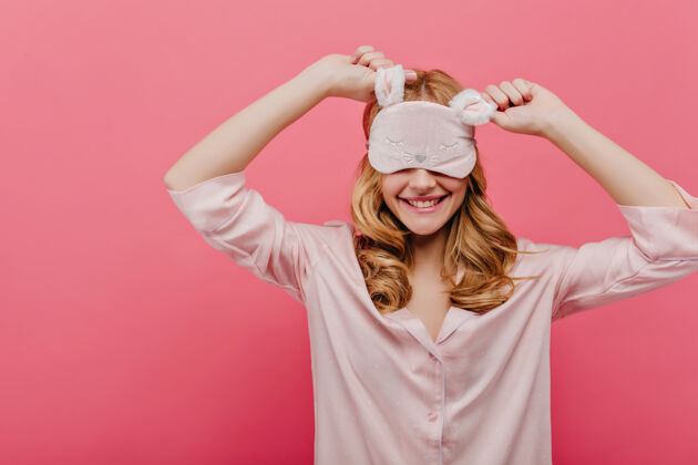 表情美丽的女孩在睡前兴高采烈迷人的卷发女人戴着眼罩和睡衣在粉红色的墙上微笑微笑女人眼罩