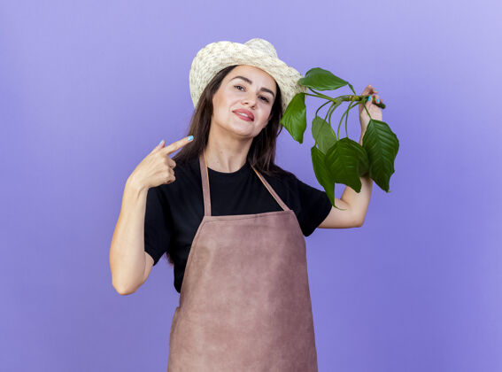 制服一个穿着制服 面带微笑的美丽园丁女孩戴着园艺帽 指着蓝色背景上孤立的植物花园帽子园艺