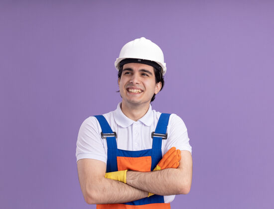 站立年轻的建筑工人身穿建筑制服 戴着安全帽 戴着橡胶手套 面带微笑 双臂交叉 站在紫色的墙上橡皮欢呼制服