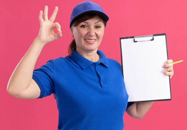 欢呼中年女送货员身穿蓝色制服 戴着帽子 拿着剪贴板 空白的书页 面带微笑地看着前面 粉红色的墙上挂着“ok”的牌子空白剪贴板制服