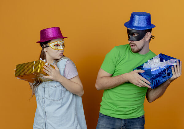 男人一对年轻贪婪的夫妇戴着粉色和蓝色的帽子 戴着化装眼罩 拿着礼盒 在橘色的墙上相互孤立地看着对方女人帽子狂欢节
