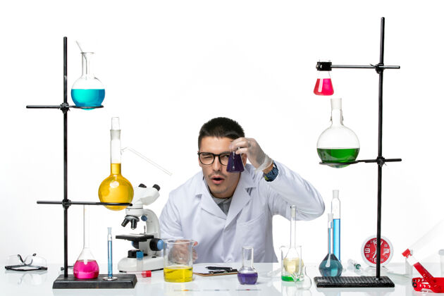 解决方案正面图身穿医疗服的男性化学家坐着拿着一个白色背景上的紫色溶液烧瓶病毒冠状病毒飞溅疾病科学科学医疗医院