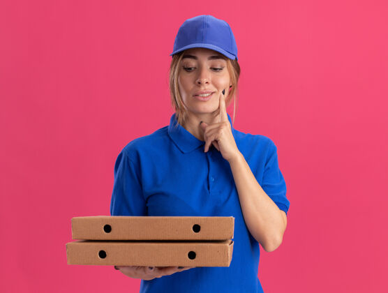 制服穿着制服的年轻漂亮的送货女孩高兴地把手指放在脸上拿着 看着粉红色的比萨饼盒披萨盒子漂亮