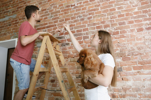 民族年轻夫妇一起修缮公寓已婚男女一起装修房子关系 家庭 宠物 爱情的概念在墙上讨论未来的设计Diy年轻油漆