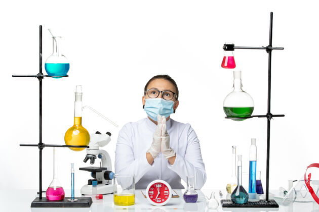实验室前视图穿着白色医疗服的女化学家带着面罩坐在浅白背景上的溶液化学家病毒covid-splashlab前面坐着瓶子