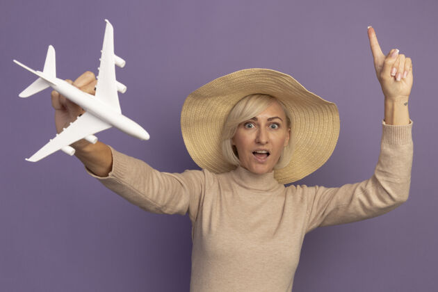 姿势一个戴着沙滩帽的漂亮的金发斯拉夫女人拿着飞机模型 指着紫色模特成人时尚