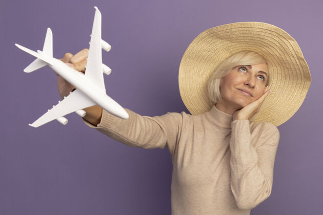 年轻漂亮的金发斯拉夫女人戴着沙滩帽 手放在脸上 手拿着飞机模型 抬头看着紫色海滩成年人时尚