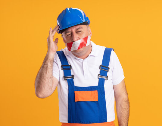 手一个穿着制服的白人成年建筑工人用胶带封住嘴巴 把手放在橘子树的头上封面人不愉快