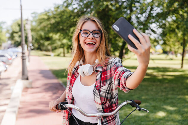 骑自行车可爱的金发女孩在公园里用智能手机自拍迷人的戴眼镜的女士骑自行车积极骑自行车田野