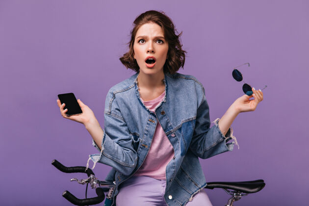 自行车忧心忡忡的黑眼睛女人拿着手机摆姿势穿着牛仔夹克的情绪激动的白人女士站在自行车旁的室内照片车辆女士美丽