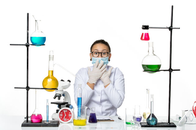 面罩前视图穿着医疗服的女化学家带着面罩坐在桌子前 白色背景上有溶液病毒化学实验室的covid飞溅物烧杯实验室研究