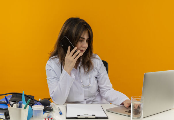成人集中中年女医生穿着医用长袍和听诊器坐在办公桌前用医用工具剪贴板用笔记本电脑讲电话橙色中年医疗