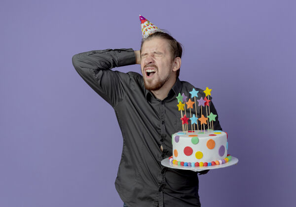 生日戴着生日帽的酸痛帅哥把头放在身后 把蛋糕孤立地放在紫色的墙上蛋糕帽子紫色