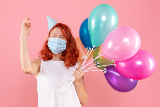 年轻女性正面图粉色书桌上戴面具手持彩色气球的年轻女性彩色病毒冠状病毒-圣诞派对庆祝正面面具