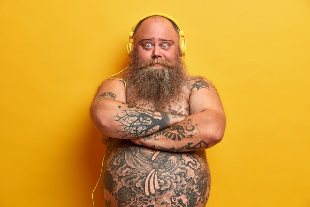 肥胖胖乎乎的男性梅洛曼双臂交叉站立 看起来很自信 身上有纹身 戴着耳机听音乐 浓密的胡须 大胖胖的肚子 隔离在黄色的墙上姿势裸露表情