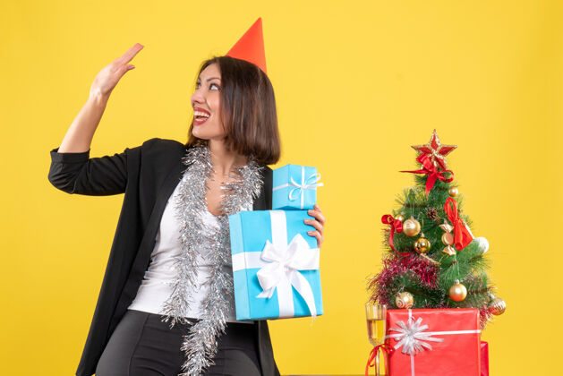 成人圣诞心情带着微笑的美丽女士戴着圣诞帽拿着礼物在黄色的办公室欢迎帽子美女帽子