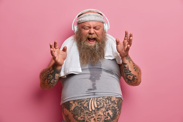 饮食情绪化的胖乎乎的潮人戴着耳机听音乐 高声唱歌 穿着运动服 进行减肥健身训练 对着玫瑰色的墙壁摆姿势运动员大胡子男人在室内烦躁的超重头带