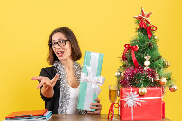 什么一位身着西装 戴着眼镜 微笑着展示礼物的商务女士 坐在办公室的一张桌子旁 桌上放着一棵圣诞树生活导演问