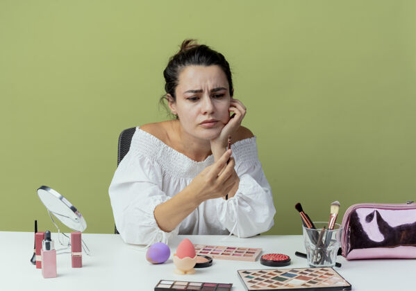 女人恼怒的美女拿着化妆工具坐在桌边 看着隔离在绿墙上的化妆刷桌子化妆坐着