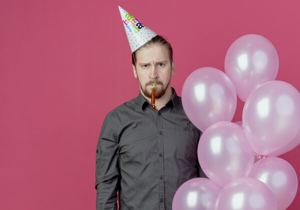 吹戴着生日帽的恼怒帅哥站在粉红色的墙上吹着氦气球粉色哨子生日