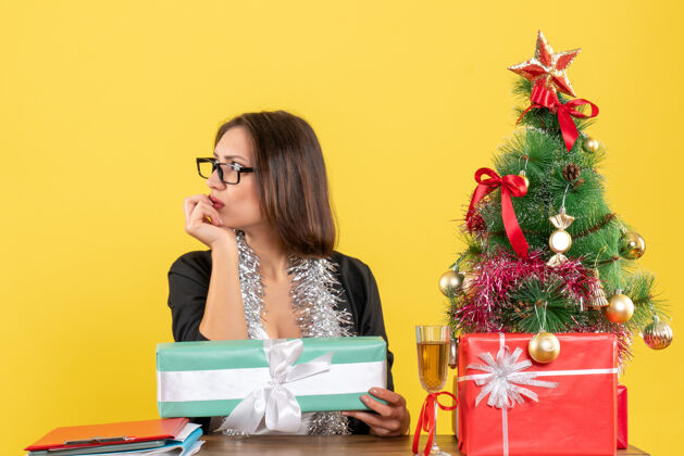 漂亮一位穿着西装 戴着眼镜 沉思着的商务女士指着她的礼物 坐在一张桌子旁 桌子上放着一棵圣诞树导演树思想