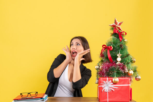 桌子震惊的年轻女子坐在一张桌子旁看着什么东西装饰圣诞树在黄色办公室树娱乐性感