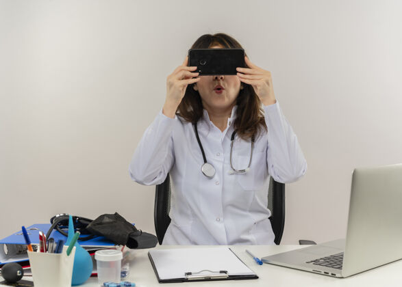 听诊器印象深刻的中年女医生穿着医用长袍和听诊器坐在办公桌前 拿着医疗工具剪贴板和笔记本电脑 躲在手机后面与世隔绝中年长袍成人